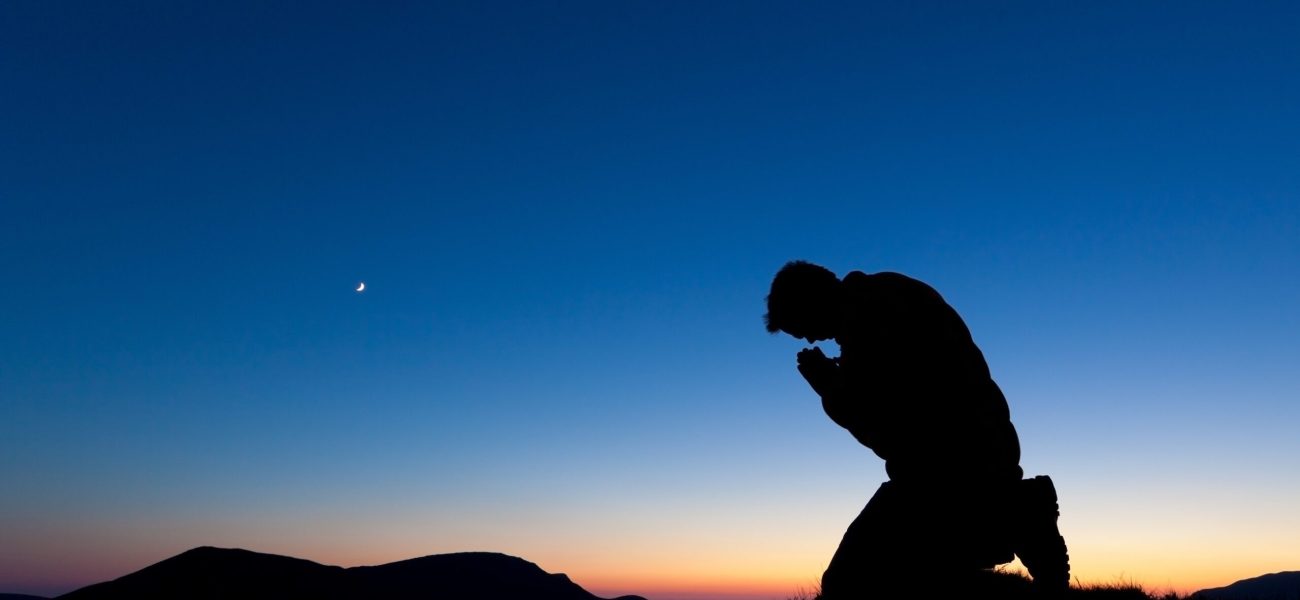 Man praying on the summit of a mountain at sun set with the moon in the sky.