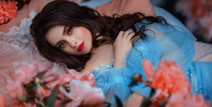 Portrait fantasy woman sleeping beauty lies on comfortable bed soft pillow. Background mystical garden, pink peonies flowers green trees. Fairy-tale girl princess cute face red lips makeup, blue dress