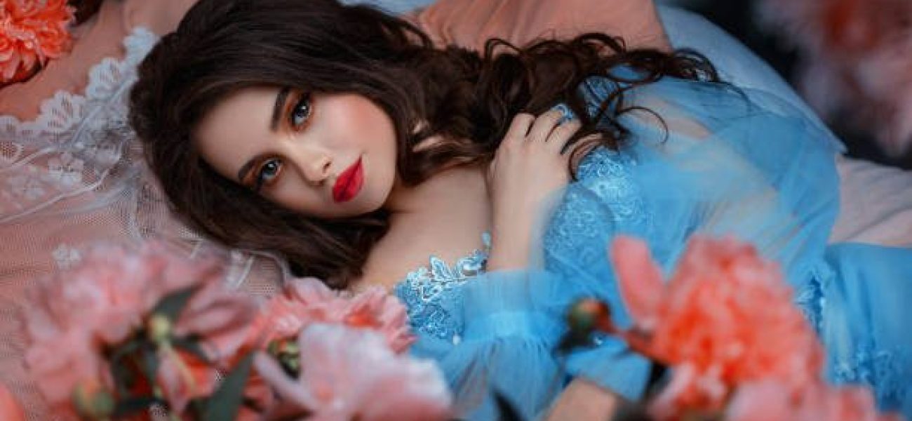 Portrait fantasy woman sleeping beauty lies on comfortable bed soft pillow. Background mystical garden, pink peonies flowers green trees. Fairy-tale girl princess cute face red lips makeup, blue dress
